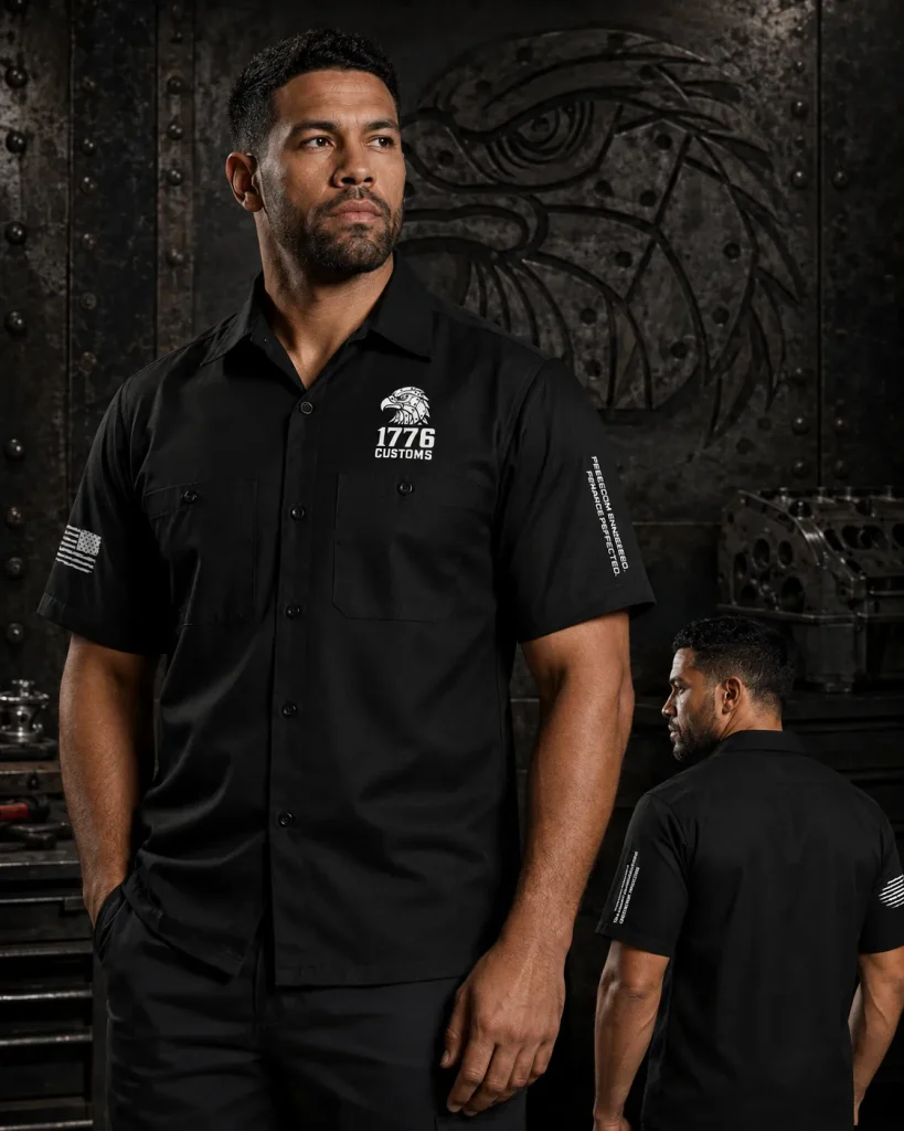 Front and back view of a male model wearing the 1776 Customs black short-sleeve button-up work shirt featuring an embroidered eagle logo on the chest, an American flag patch on the sleeve, and vertical brand text on the arm against an industrial dark metal backdrop.
